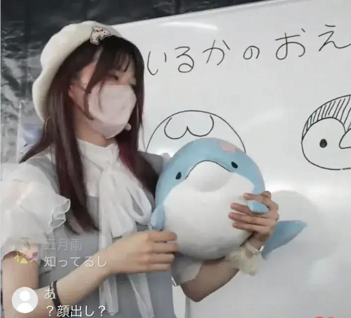 みいるかが教室のような場所でイルカのぬいぐるみを抱え、白いベレー帽とマスク姿で立っている写真